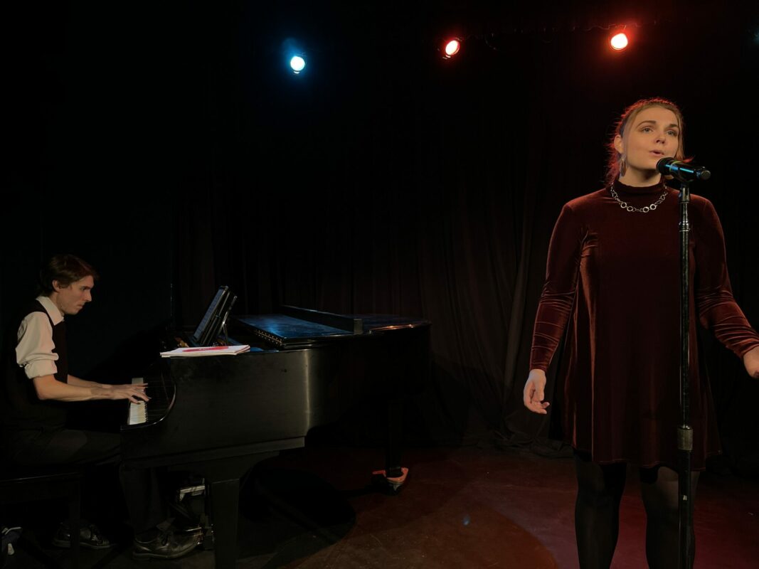 Woman singing into a microphone while a man wearing a vest plays the piano.