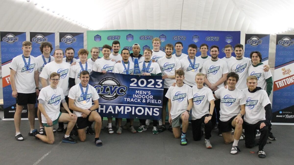 Men's track team wearing matching shirts with some wearing medals around their neck. In the center, there is a banner that reads "2023 Men's Indoor track and field champions"
