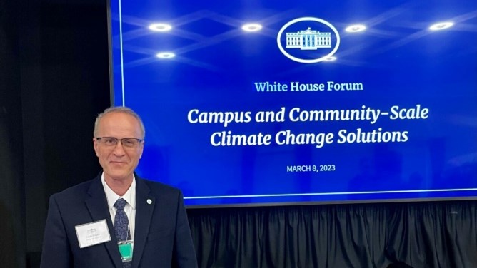 Man standing in front of a screen that says "White House Forum" "Campus and Community-Scale Climate Change Solutions"