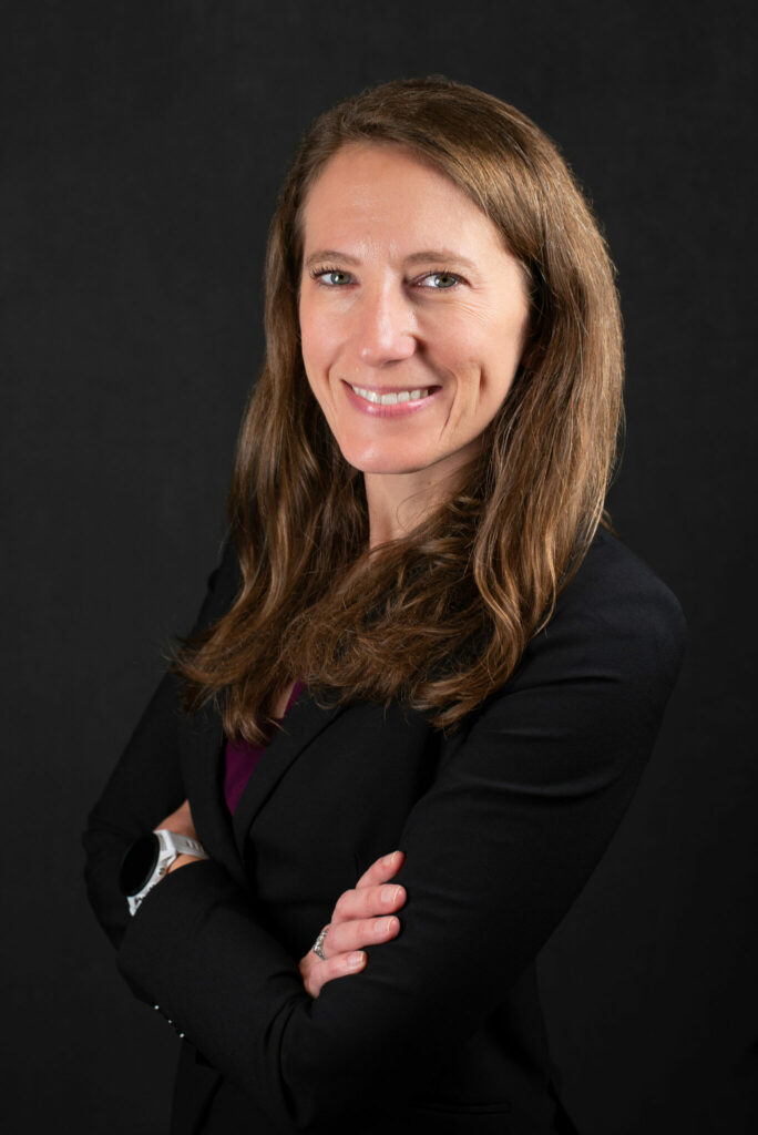 Portrait of Dr. Kate Sheppard standing with her arms crossed, wearing a black blazer over a dark top, photographed against a dark studio background.