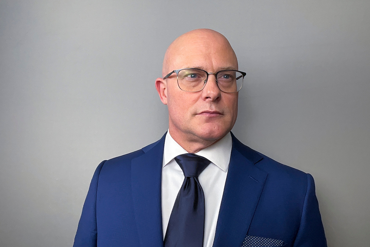 Person in a blue suit and tie wearing glasses, standing against a plain gray background and looking slightly to the side.
