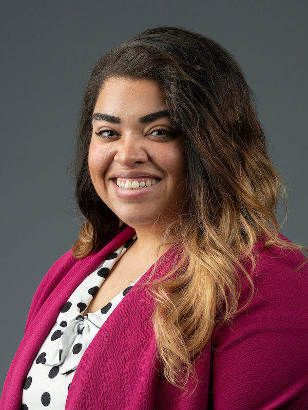 Smiling woman with long, wavy hair wearing a magenta blazer and polka dot blouse, set against a neutral gray background.