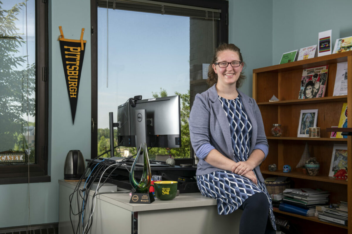 A person sitting on a desk in an office, smiling. Behind them, a wooden shelf with books and art, and a "Pittsburgh" pennant on the wall.