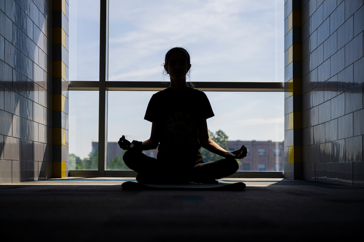 Silhouette of a person meditating in a lotus position indoors, backlit by sunlight through a large window, creating a serene and peaceful ambiance.