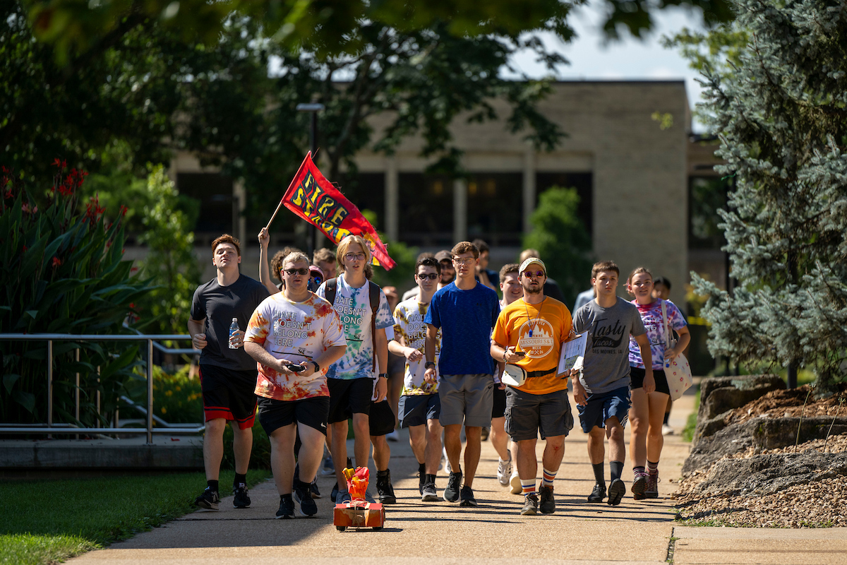 A group of young adults walk on a sunny campus path, carrying a red and yellow flag following an RC car. They exude a lively and energetic atmosphere.