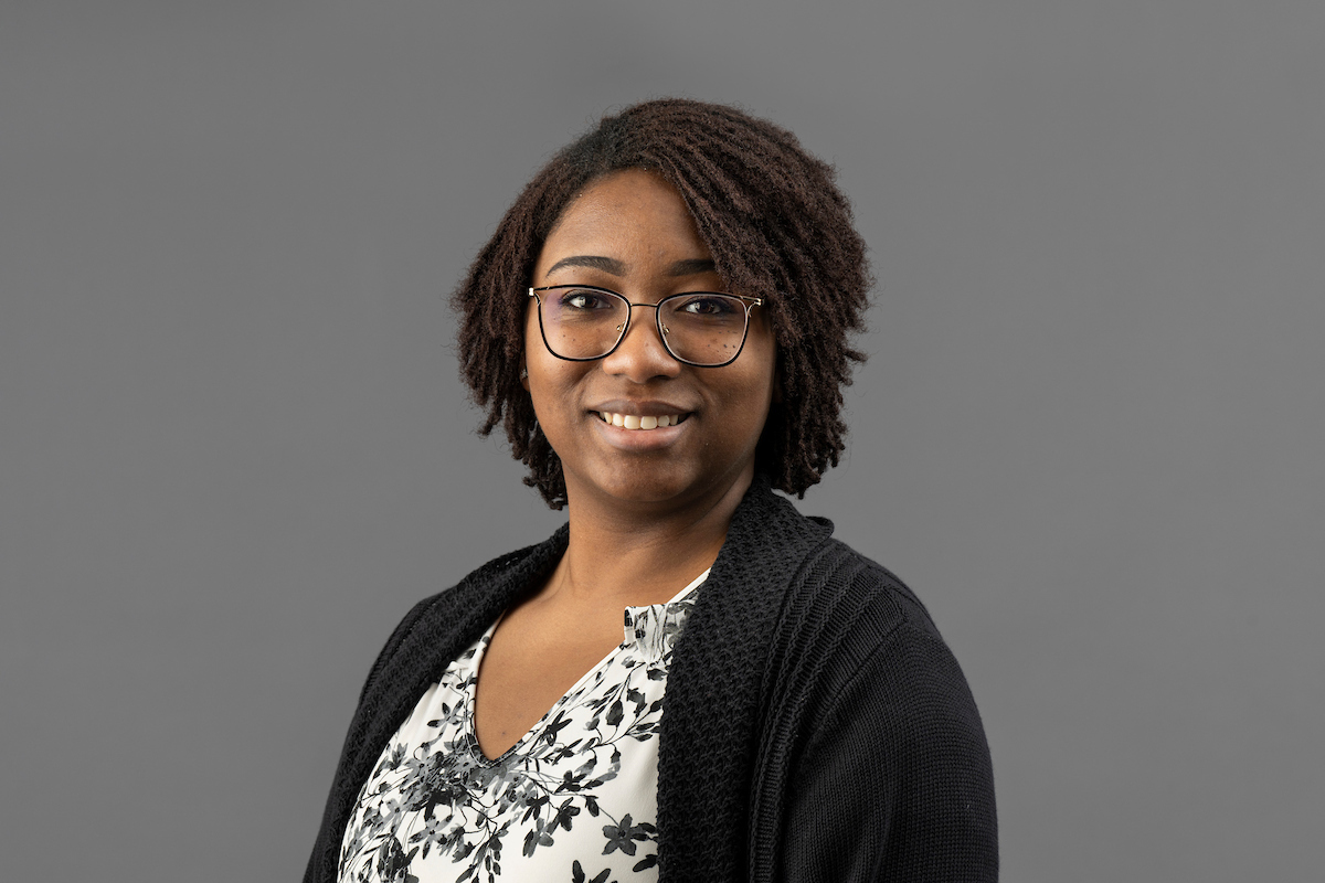Smiling woman with glasses, wearing a floral blouse and dark cardigan, against a plain gray background.