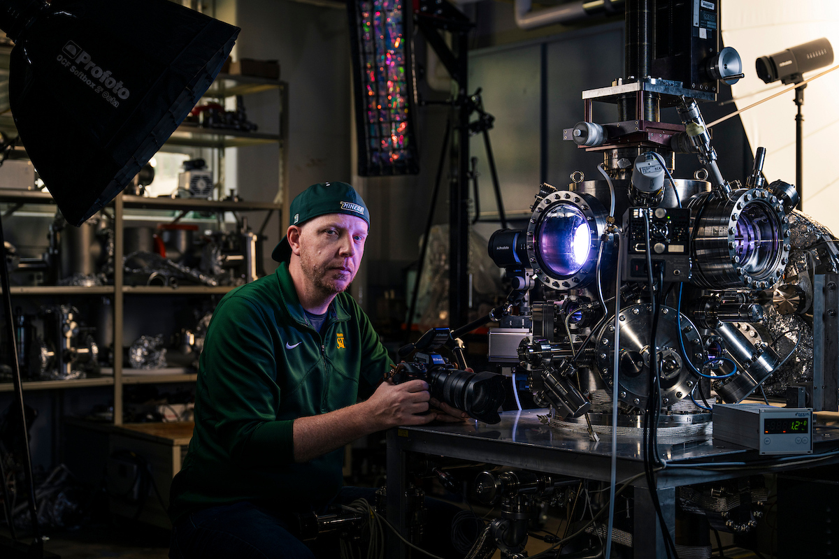 Man in green sports attire and cap holding a camera, sitting beside complex machinery emitting blue light.