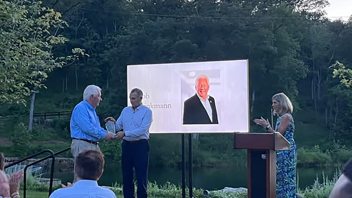 A man receives an award on an outdoor stage with trees in the background. A large screen shows a portrait of the man receiving the award