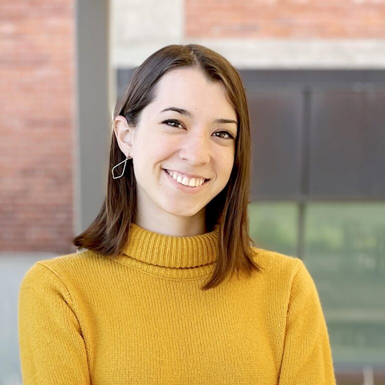 Young woman with shoulder-length brown hair, smiling, wears a yellow sweater and geometric earrings.