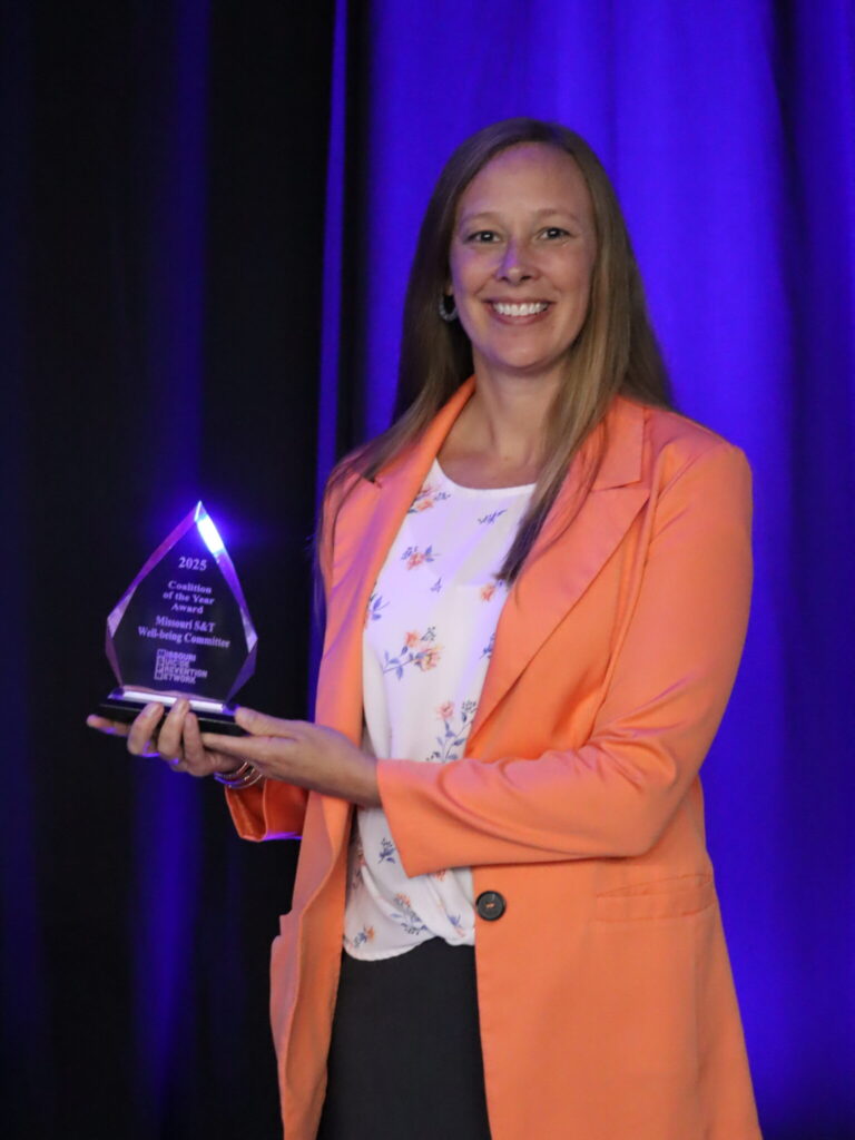 A woman smiling wearing an orange blazer with a floral top holding an award.