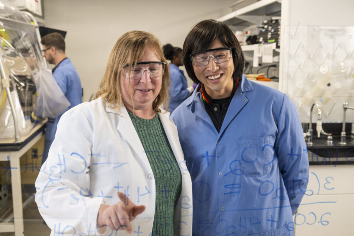 Two scientists in lab coats smile while working on chemical equations on a transparent board. The laboratory setting is busy, with others working in the background.