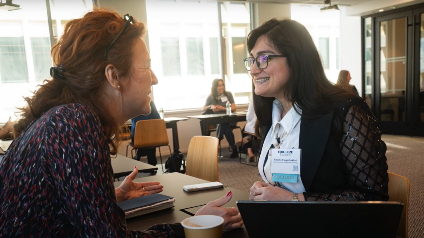 Two women smiling and talking at a conference in a bright room with large windows. They sit at a table with papers, a phone, and a coffee cup.