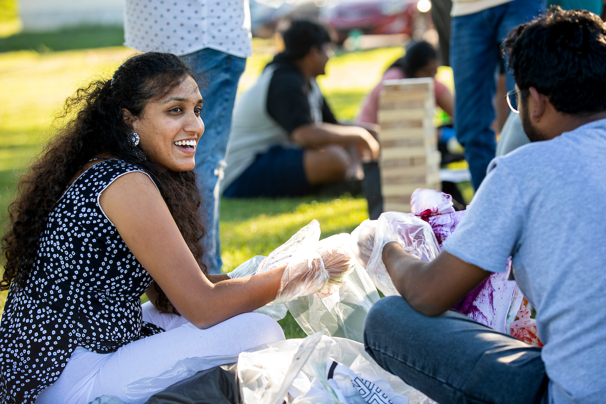 Two students sit on the ground to tie dye T-shirts while others play a game of Jenga in the background.