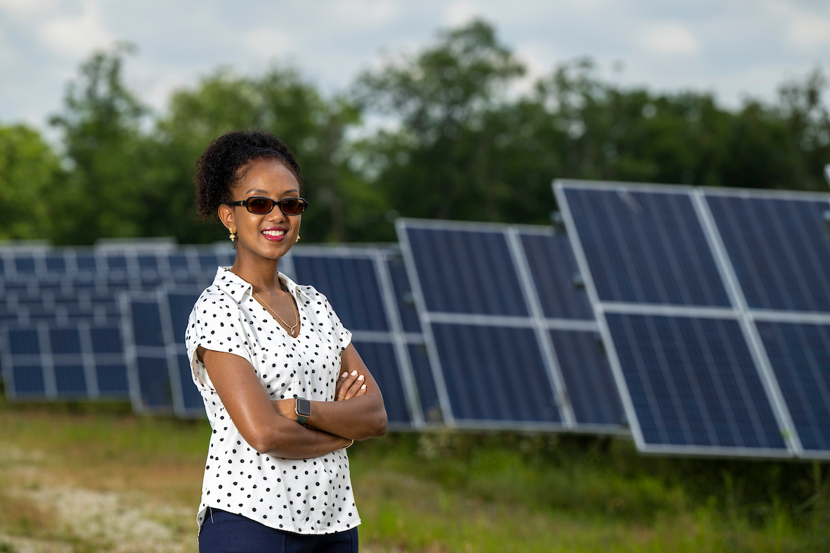 A woman stands confidently with crossed arms in front of a field of solar panels. She wears sunglasses and a polka-dot blouse.
