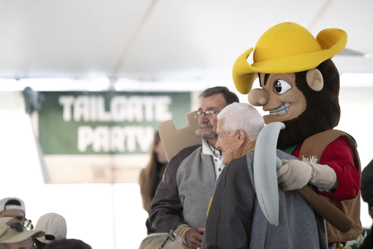 Joe Miner, S&T's mascot, poses with two older adults in front of a "Tailgate Party" sign.
