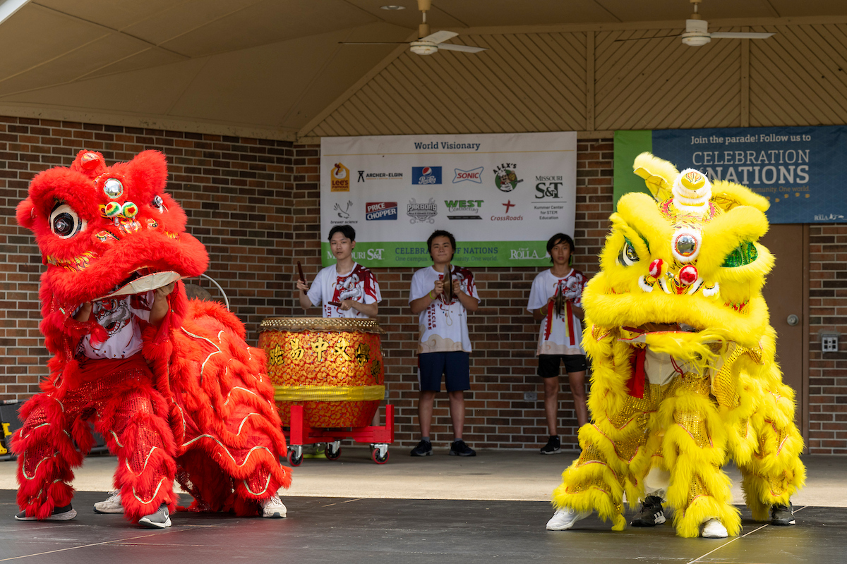 Two performers in colorful costumes on stage during a cultural performance at the 2024 Celebration of Nations festival, with musicians and banners in the background.