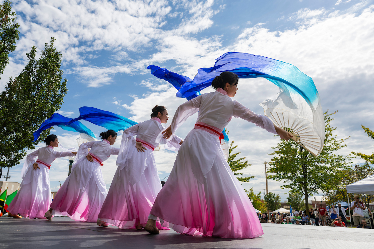 Four women dancing on a stage wearing white and pink dresses with fans with blue fabric attached.