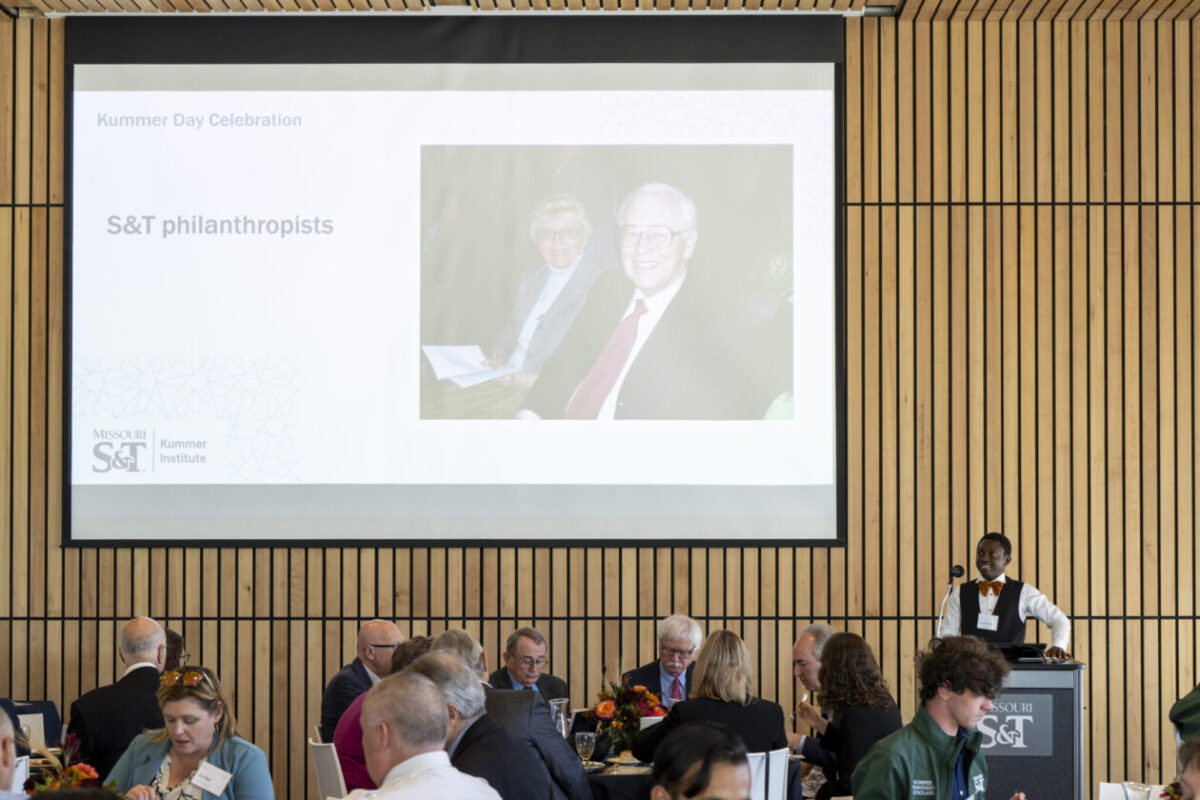 A student stands at a podium in front of the room near a projector with tables of people.