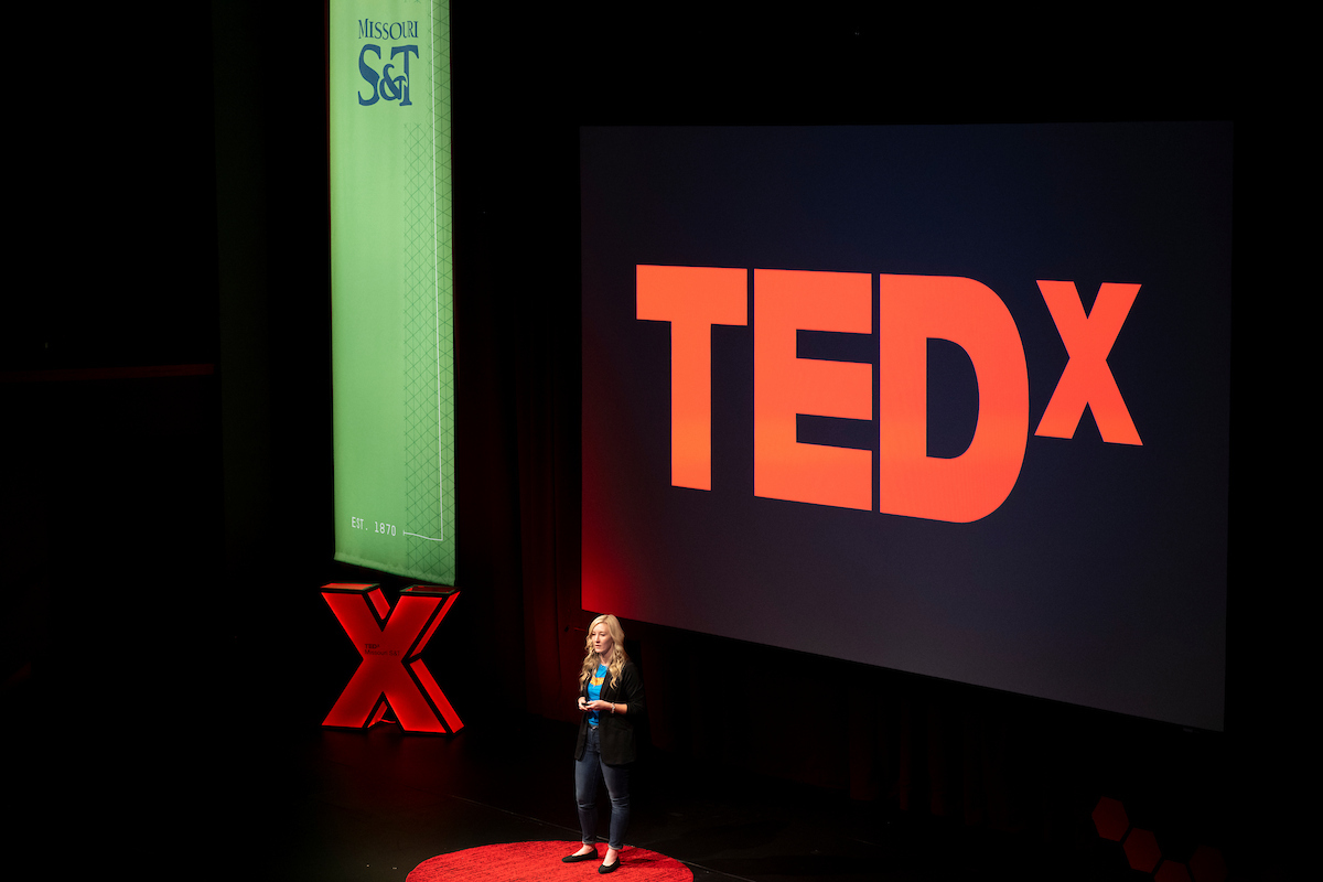 Woman on a stage standing in a red circle and in front of a projector that says TEDx.