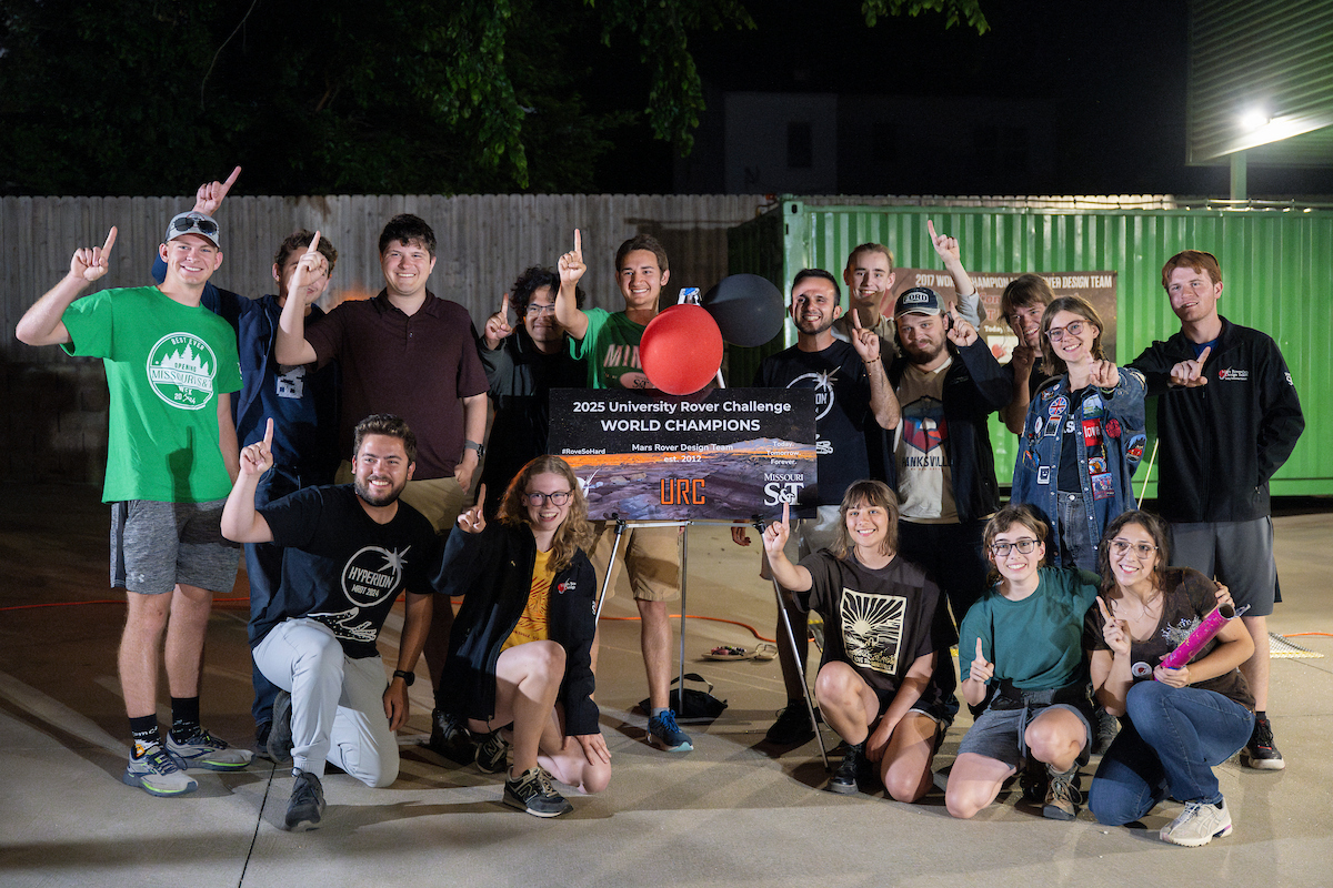 A group of people celebrate winning the 2023 University Rover Challenge, holding a sign that reads "World Champions." They are smiling and pointing upwards, conveying excitement and achievement.