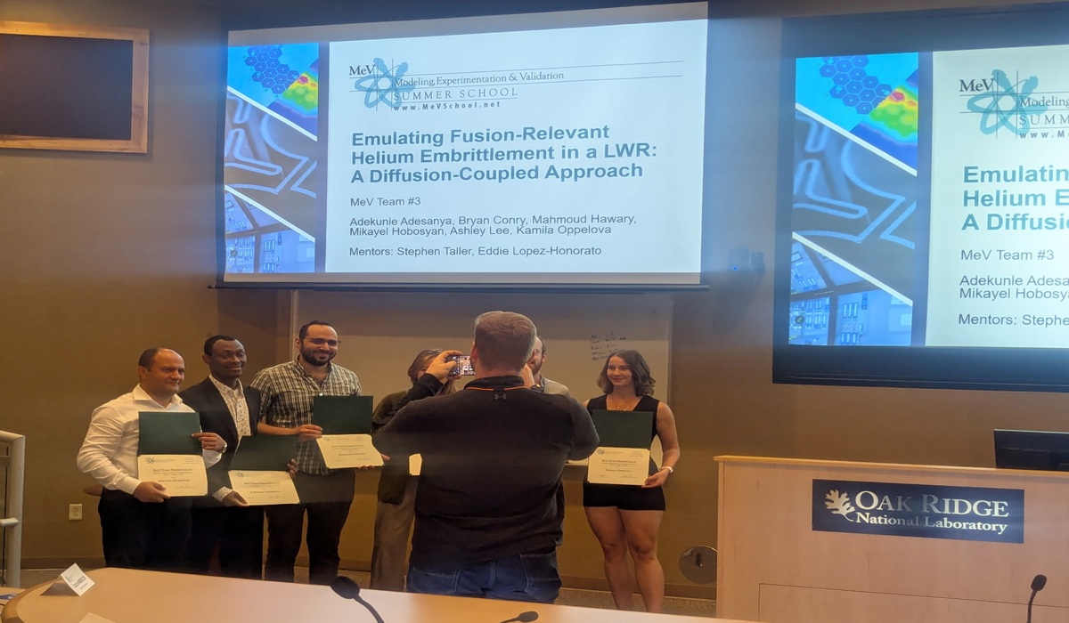 A group of six people stands in front of a large projection screen holding certificates, smiling for a photo at Oak Ridge National Laboratory.
