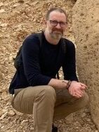 A man in a black long sleeve shirt and brown pants with glasses, kneels next to a rock.