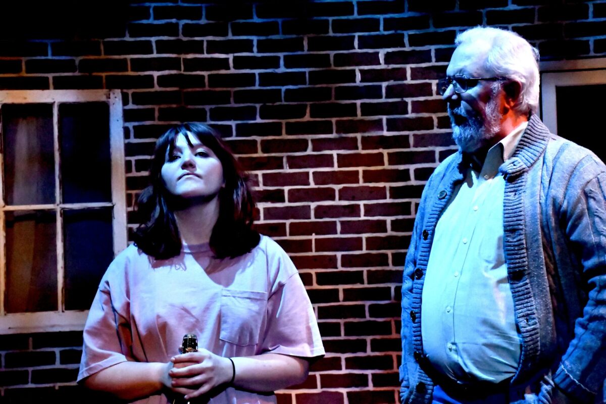 A young woman with dark hair stands holding a bottle, looking contemplative. An older man in a cardigan stands beside her, both against a brick wall.
