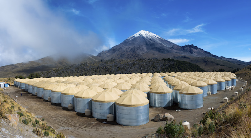 An array of covered corrugated steel tanks in front of a snow-covered mountain.