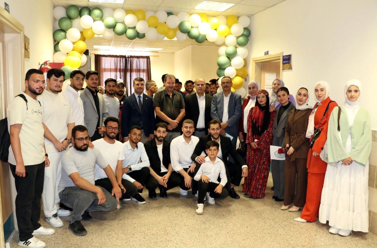 A diverse group of people stand in a hallway under an archway of green, yellow, and white balloons. They are dressed in formal and cultural attire, smiling warmly at the camera, conveying a celebratory and joyful atmosphere.