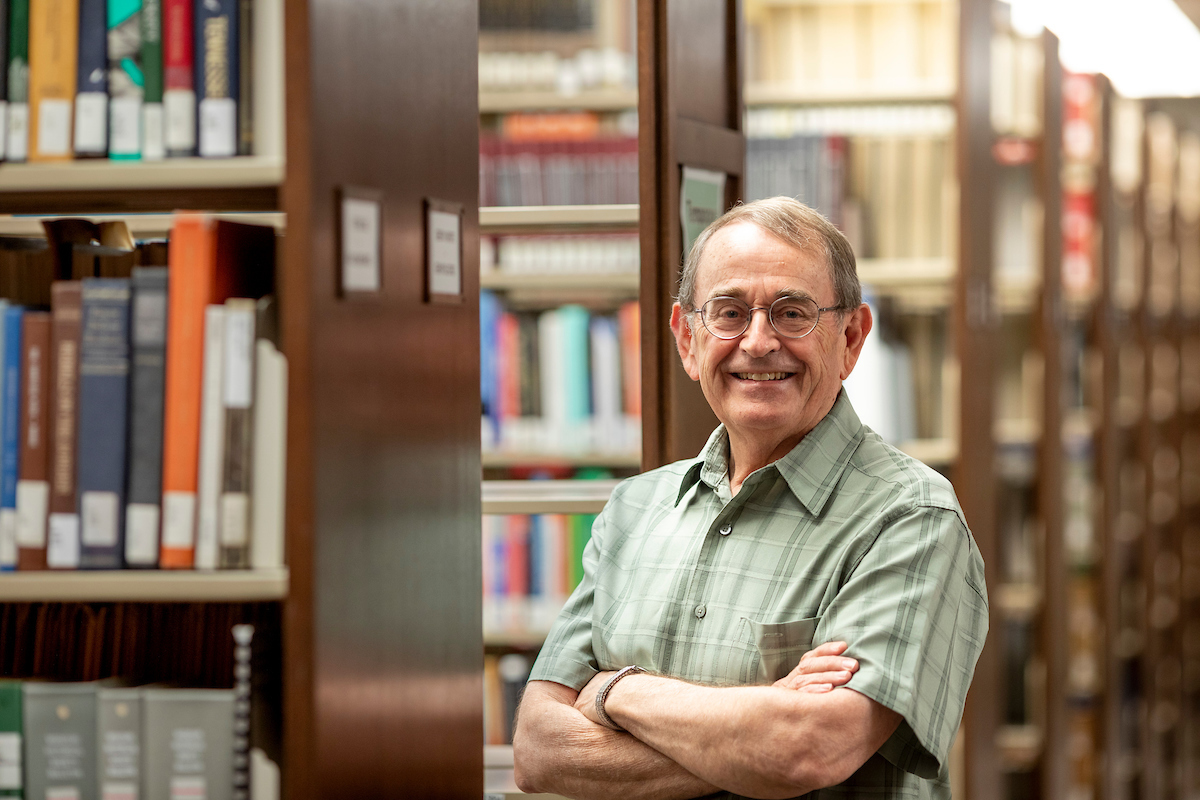 Dr. Gragg stands with his arms crossed and a smile near bookshelves in a library.