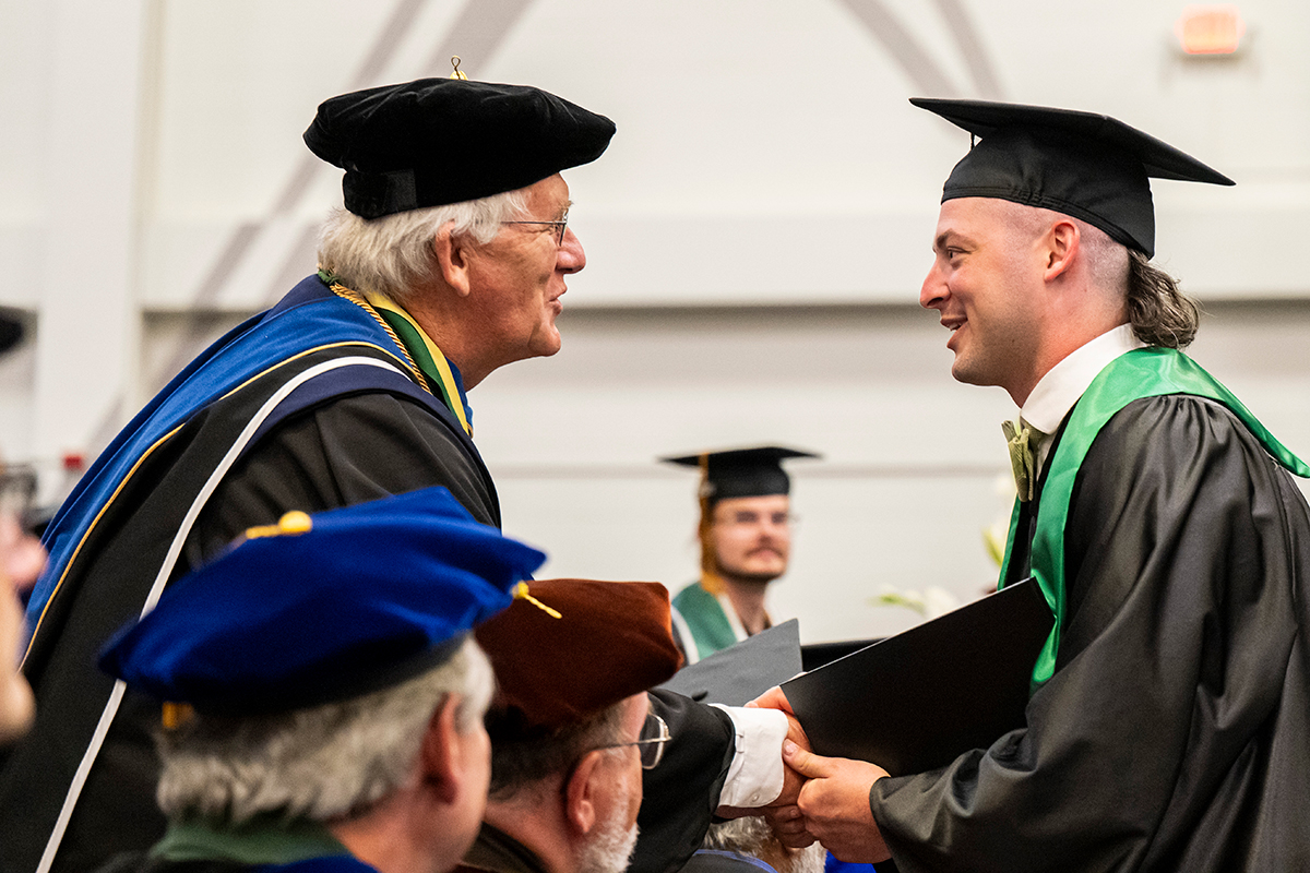 A faculty member shakes the hand of a student while wearing commencement regalia.
