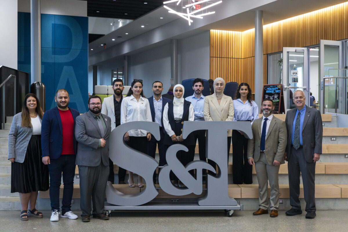 Twelve people take a photo with a large monument in the shape of S&T.