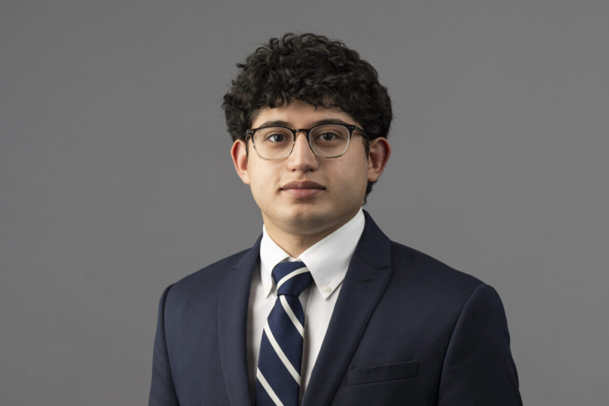 A student wearing a jacket and striped tie and glasses with a neutral facial expression.