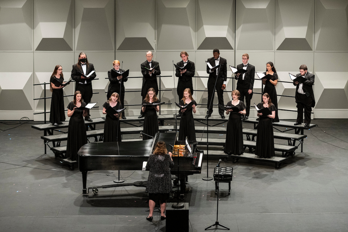 A choir stands on risers as they perform a concert with a director conducting.