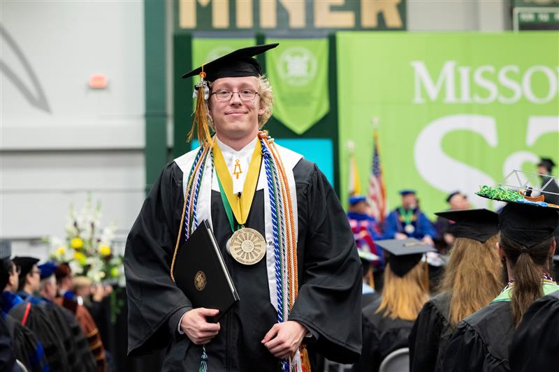 A student in graduation regalia wears many stoles and has a large medal hanging around his neck.