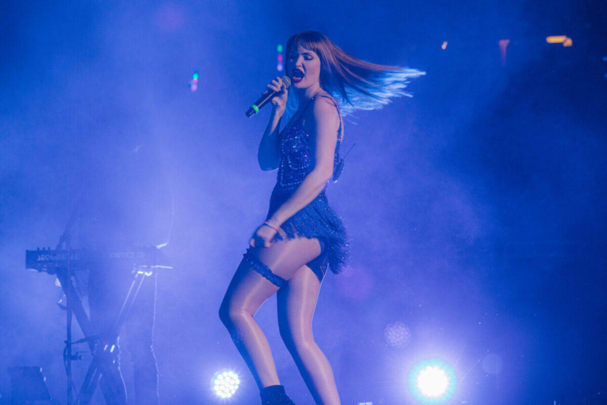A woman sings into a microphone on a stage with purple lights and a purple dress.