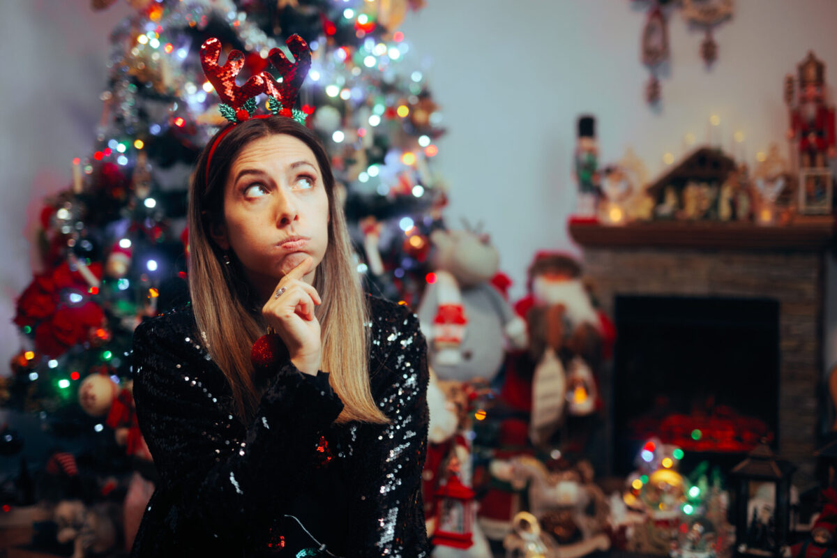 A woman sits in front of a Christmas tree looking perplexed while wearing a headband with red shiny antlers.