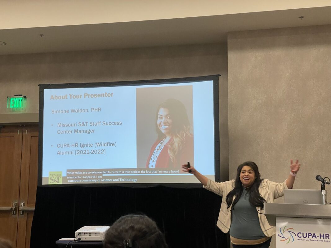 Simone giving a presentation in front of a projector screen with her hands in the air with excitement.