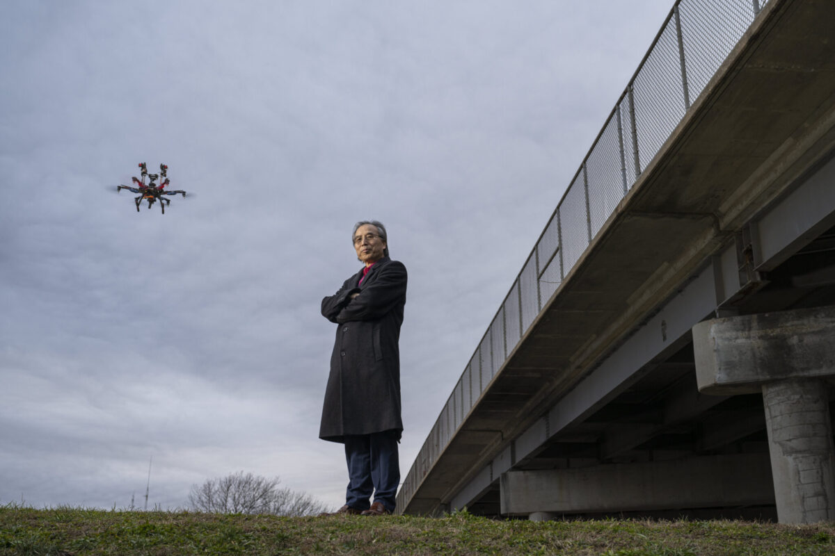 A man standing near a bridge wearing a trench coat with a drone flying over head.