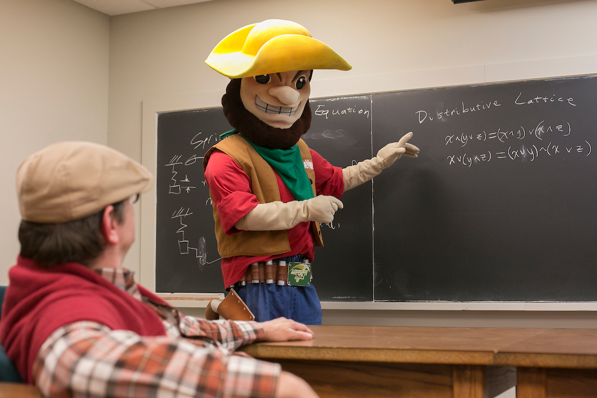 A person wearing the Joe Miner mascot costume points to a black board with math equations.
