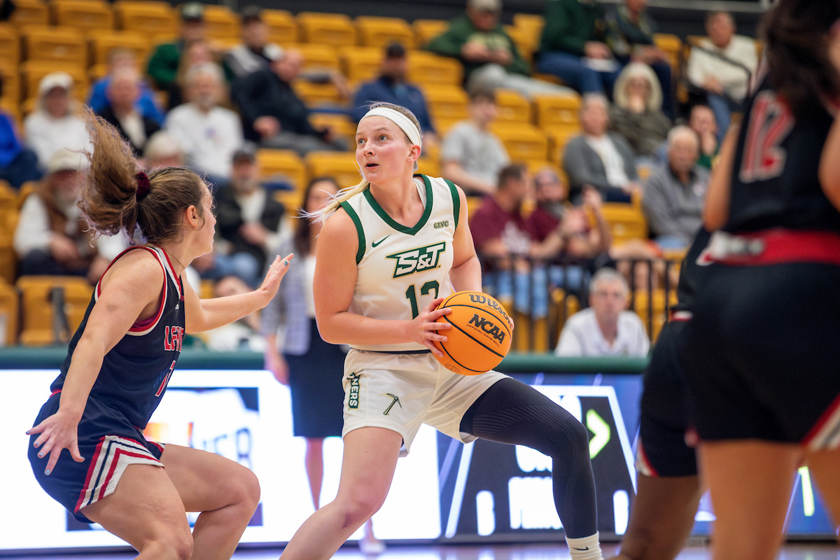 A woman with a Miner's basketball jersey holds the basketball while looking for an opening to shoot.