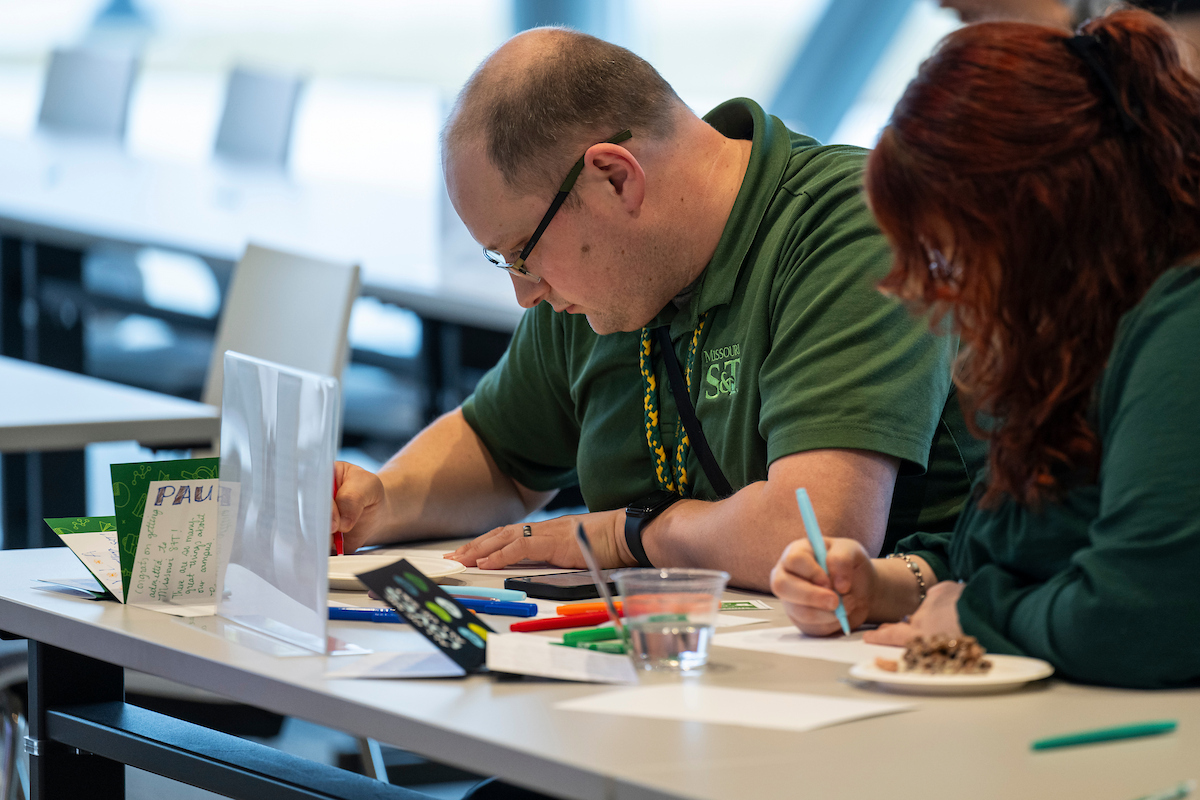 A man wearing an S&T green polo and a woman with red hair write in cards with colorful markers.