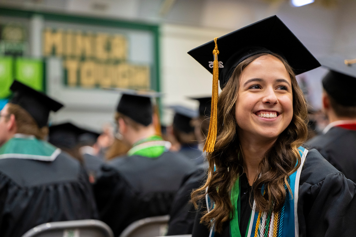 A woman, wearing a graduation cap with a gold tassel and gown with several stoles and cords, smiles to family and friends attending the commencement event.