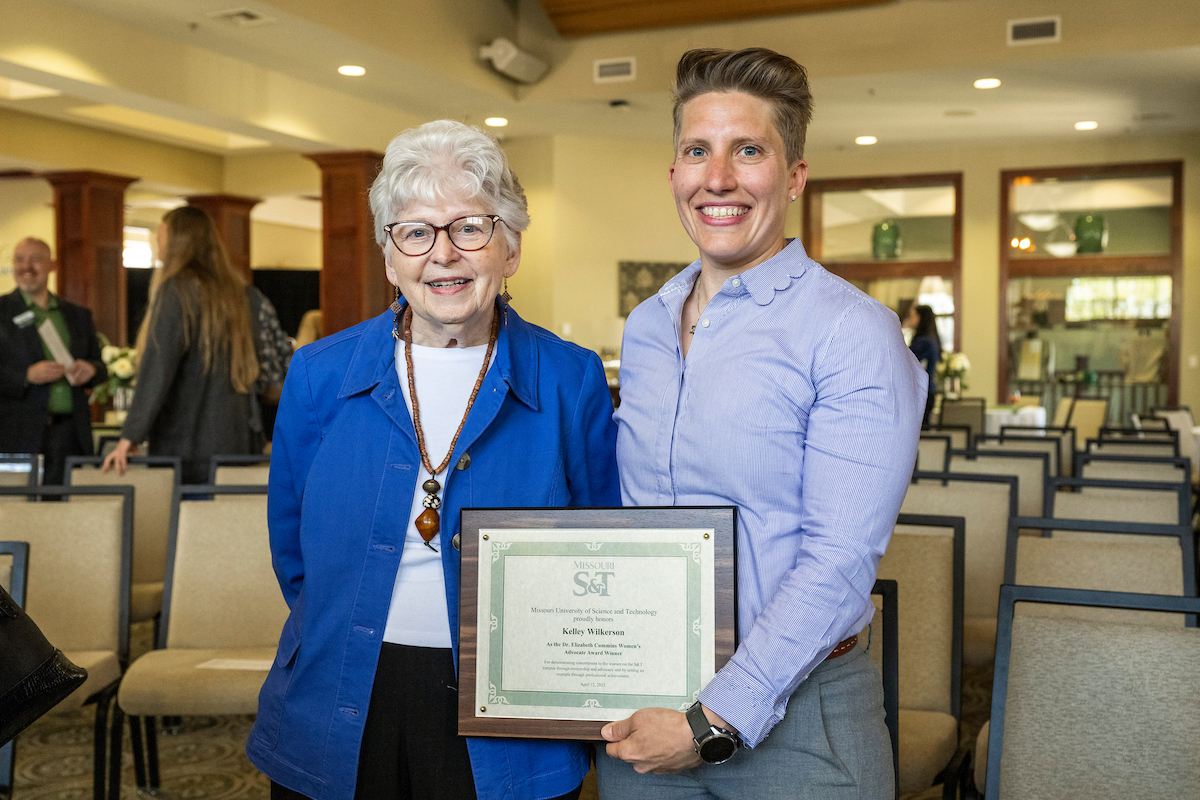 Two smiling individuals stand in a bright room with rows of empty chairs. One holds a framed certificate, conveying a sense of achievement and celebration.