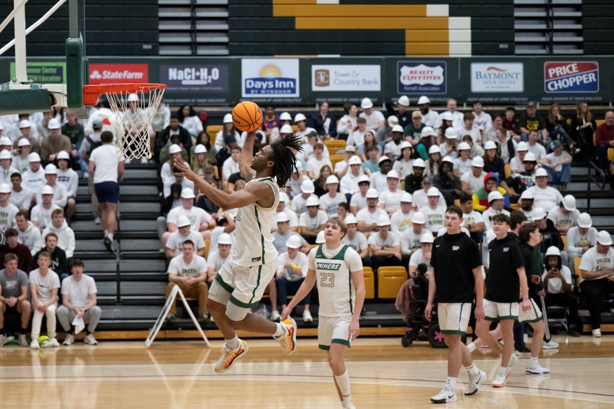 A man dressed in a white and green uniform elevates toward the basketball goal for a right handed lay up.