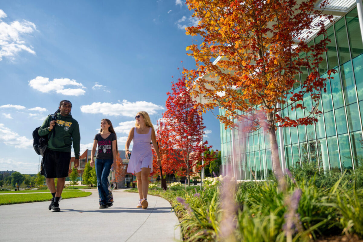 Three students walk along the sidewalk in front of the Welcome Center.