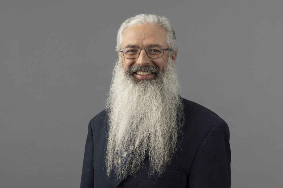 Studio portrait of Dr. Jonathan Kimball with long white hair and a full white beard, wearing a dark jacket, photographed against a neutral gray background.