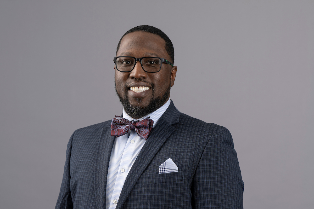 Man wearing dark glasses and a plaid suit with a red bow tie, smiling confidently against a neutral gray background. Professional and approachable tone.
