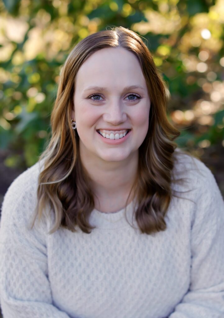 A headshot of a woman smiling with red shoulder length hair wearing a cream colored sweater.