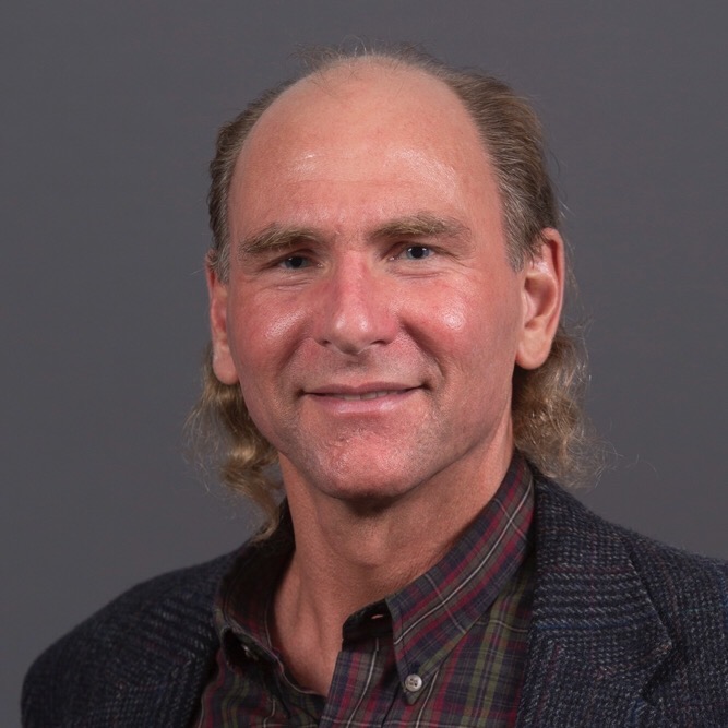 Portrait of Dr. Ross Reed, a lecturer in arts, languages, and philosophy at Missouri S&T, wearing a textured dark blazer over a plaid button-down shirt against a neutral gray background.